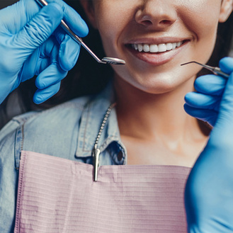 Patient in dental chair with pink bib, dentist’s gloved hands using mirror and probe near clean teeth.
